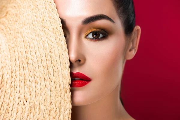 Hermosa mujer joven sosteniendo sombrero de paja y mirando a la cámara aislada en rojo - foto de stock