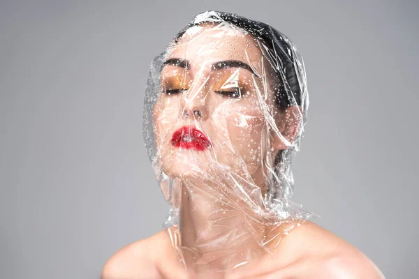 Jeune femme sensuelle avec cellophane humide transparent sur la tête et les yeux fermés isolés sur le gris — Photo de stock