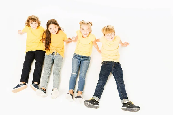 Blick aus der Vogelperspektive auf entzückende glückliche Kinder in gelben T-Shirts, die zusammen sitzen und vereinzelt in die Kamera lächeln — Stockfoto