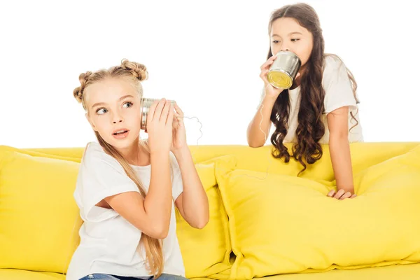 Excited kids playing with tin cans phone on yellow sofa isolated on white — Stock Photo