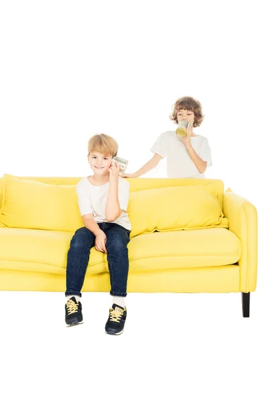 Fröhliche Jungen spielen mit Blechdosen-Telefon auf gelbem Sofa isoliert auf weißem — Stockfoto