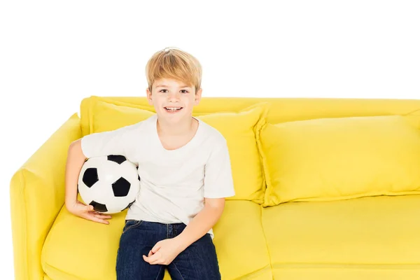 Улыбающийся очаровательный мальчик сидит с футбольным мячом на желтом диване изолирован на белом и смотрит в камеру — стоковое фото