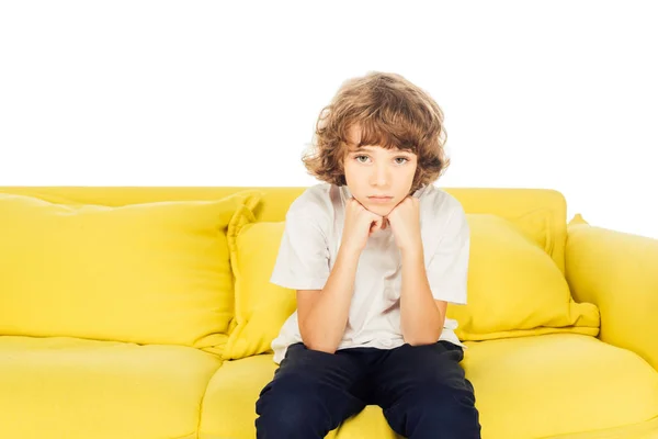 Chateado menino sentado no sofá amarelo e descansando queixo em mãos isoladas no branco, olhando para a câmera — Fotografia de Stock