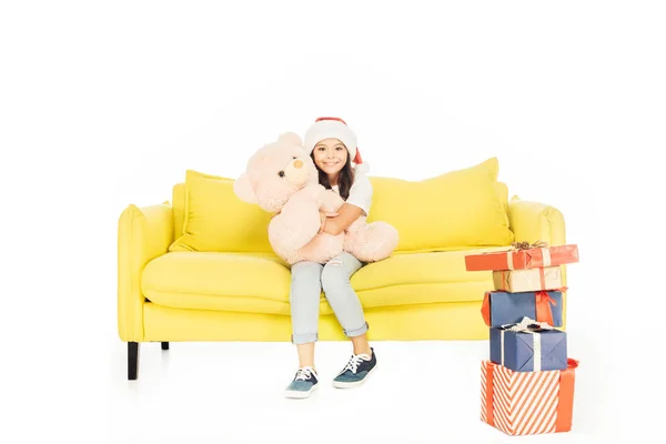 Adorável criança em santa chapéu abraçando ursinho de pelúcia no sofá amarelo e olhando para a câmera isolada no branco — Fotografia de Stock