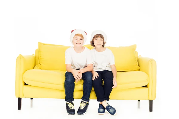 Adorable chicos en santa sombreros sentado en amarillo sofá aislado en blanco, mirando a la cámara - foto de stock