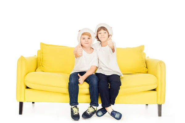 Adorables garçons en chapeaux de Père Noël assis sur canapé jaune isolé sur blanc et montrant pouces vers le haut — Photo de stock