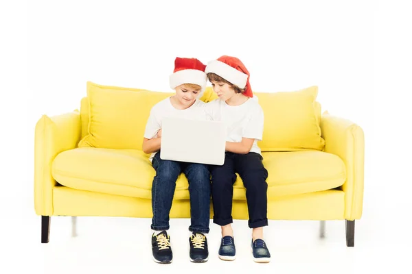 Прелестные мальчики в шляпах Санты сидят на жёлтом диване и используют ноутбук, изолированный на белом — стоковое фото