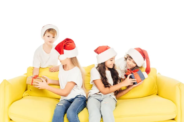 Adorabili bambini in cappelli da Babbo Natale seduti sul divano giallo con scatole regalo isolate su bianco — Foto stock