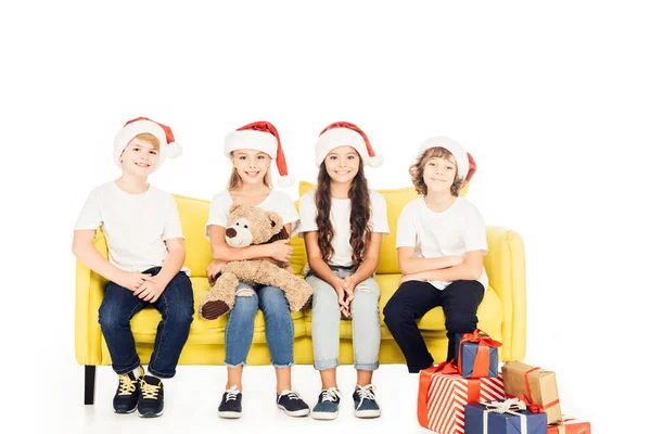 Adorable kids in santa hats sitting on yellow sofa with teddy bear and presents isolated on white — Stock Photo