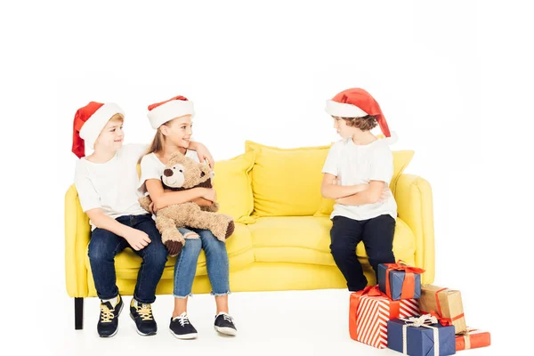 Adorable kids in santa hats hugging on yellow sofa, angry boy sitting with crossed arms isolated on white — Stock Photo