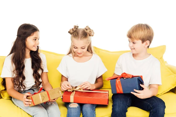 Crianças adoráveis sentadas no sofá amarelo e abrindo caixas de presente isoladas no branco — Fotografia de Stock