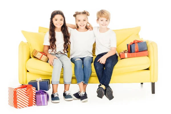 Adorables enfants assis sur un canapé jaune avec des boîtes-cadeaux isolées sur blanc — Photo de stock