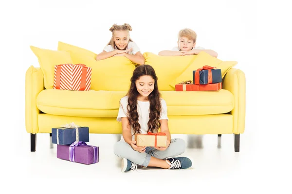 Smiling adorable child holding gift box isolated on white, friends looking out from yellow sofa — Stock Photo