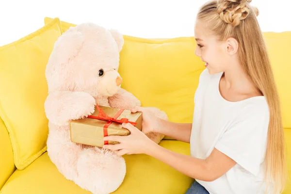 Smiling adorable child giving present to teddy bear on yellow sofa isolated on white — Stock Photo