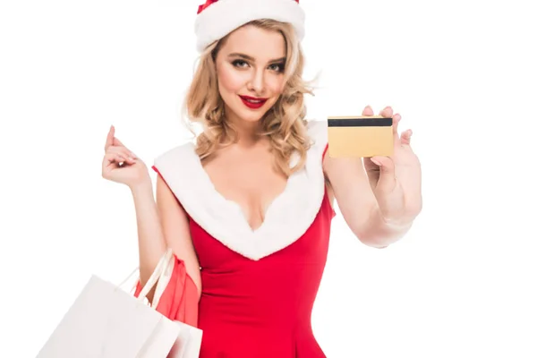 Sonriente chica santa en vestido de navidad sosteniendo bolsas de compras y mostrando la tarjeta de crédito aislado en blanco - foto de stock