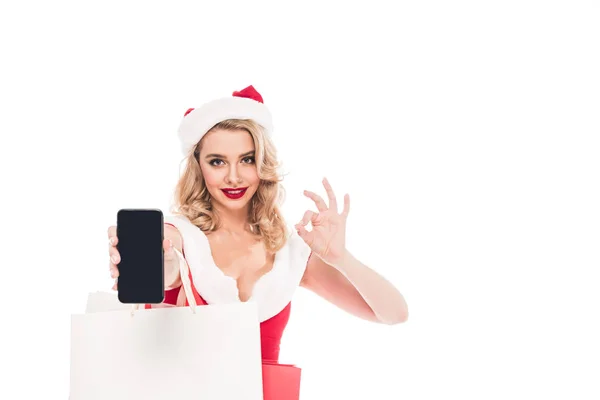 Santa fille avec des sacs à provisions faisant signe ok et montrant smartphone avec écran vide isolé sur blanc — Photo de stock