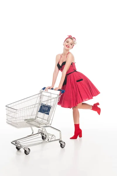 Feliz joven mujer en retro pin up vestido llevando carrito de compras aislado en blanco - foto de stock