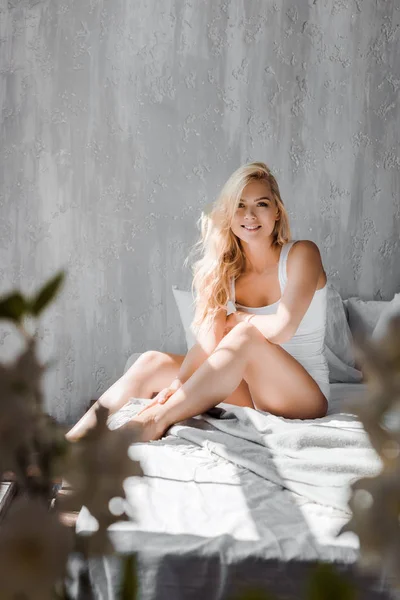 Selektiver Fokus attraktiver blonder Mädchen in Unterwäsche, die auf dem Bett sitzen und in die Kamera lächeln — Stockfoto