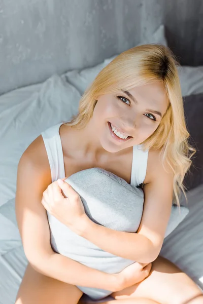 Vista ad alto angolo di felice giovane donna in biancheria intima seduta con cuscino sul letto e sorridente alla macchina fotografica — Foto stock