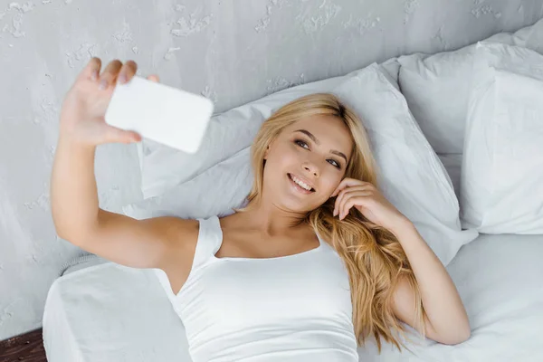 Vista ad alto angolo di sorridere giovane donna sdraiata sul letto e scattare selfie da smartphone — Foto stock