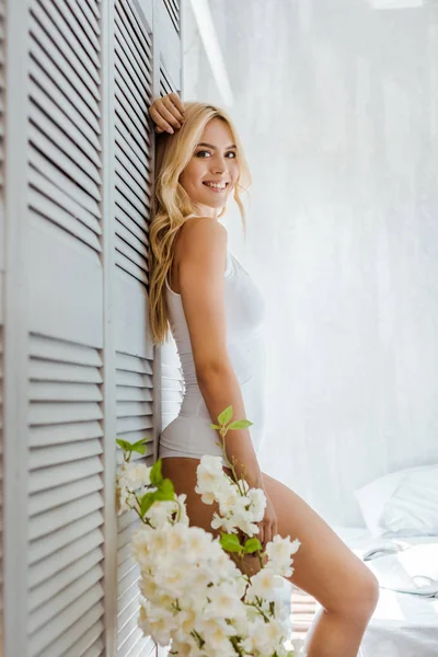 Vista lateral de hermosa chica rubia en ropa interior blanca sonriendo a la cámara en el dormitorio - foto de stock