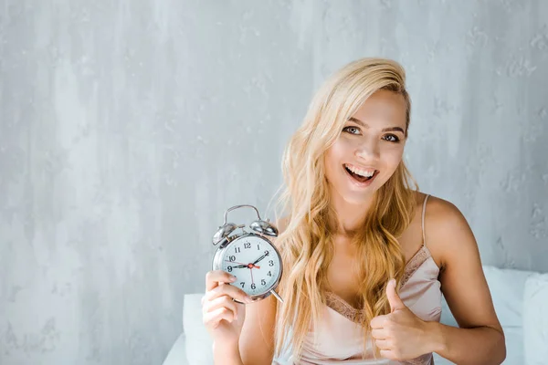 Glückliche junge Frau hält Wecker und lächelt in die Kamera im Schlafzimmer — Stockfoto