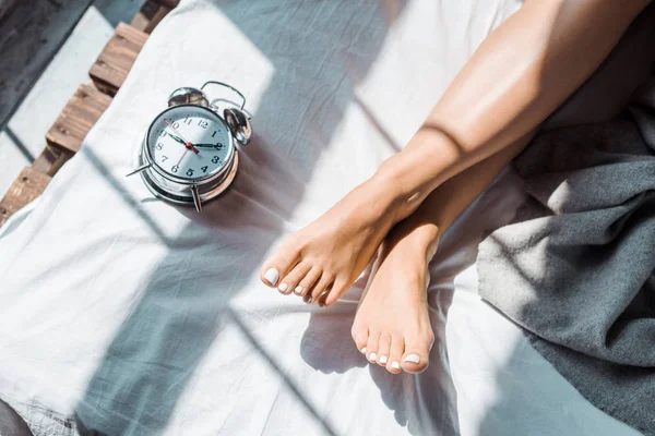 Partial top view of female feet and alarm clock on bed — Stock Photo