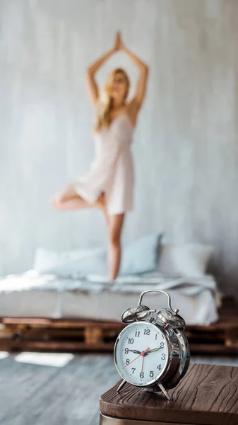Vista de cerca del reloj despertador en la mesa de madera y la joven realizando yoga en la cama detrás - foto de stock