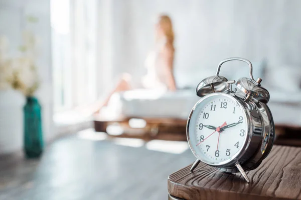 Close-up view of alarm clock on wooden table and young woman sitting on bed behind — Stock Photo
