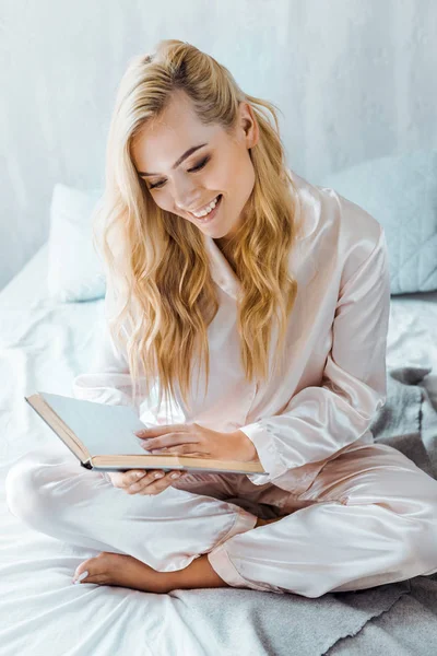 Hermosa mujer joven feliz en pijama libro de lectura en el dormitorio - foto de stock