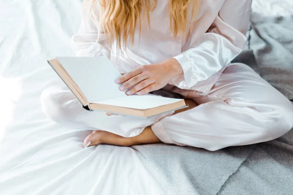 Tiro cortado de jovem mulher de pijama sentado na cama e livro de leitura — Fotografia de Stock