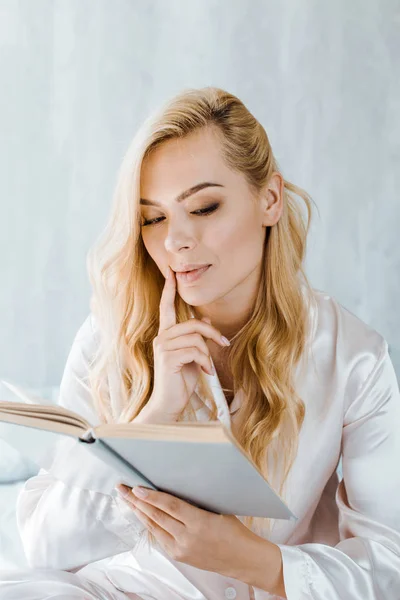 Fokussierte junge blonde Frau im Schlafanzug liest Buch im Schlafzimmer — Stockfoto