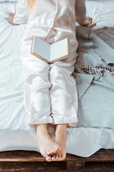 Cropped shot of young woman in pajamas sitting on bed with book — Stock Photo