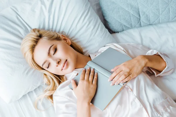 Top view of young woman in pajamas holding book and sleeping on bed — Stock Photo