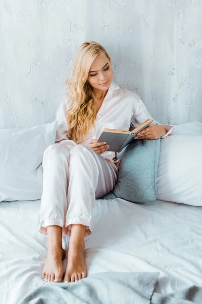 Высокий угол зрения на красивую молодую женщину в пижаме, сидящую на кровати и читающую книгу — стоковое фото