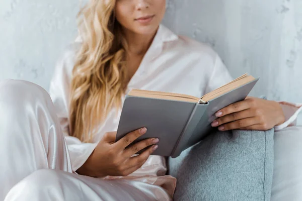 Recortado disparo de mujer joven en pijama sentado en la cama y lectura de libro - foto de stock