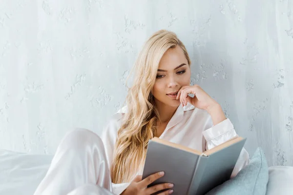 Focalizzata giovane donna in pigiama seduta sul letto e libro di lettura — Foto stock