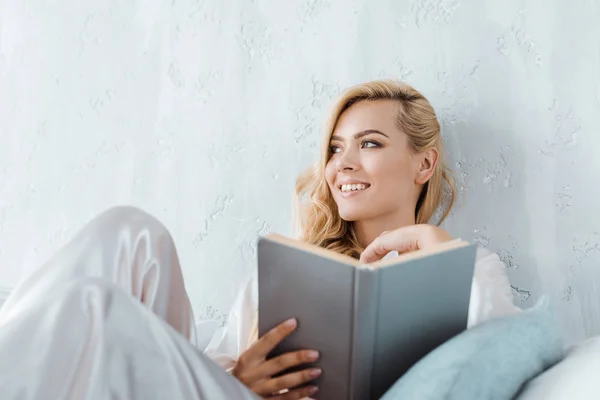 Attractive smiling young woman in pajamas holding book and looking away while in bedroom — Stock Photo