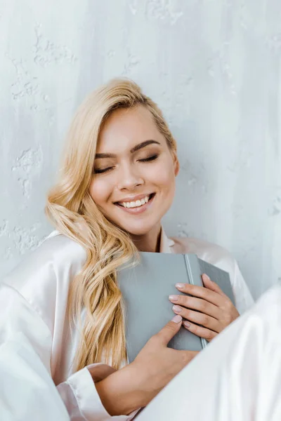 Schöne glückliche blonde Mädchen im Schlafanzug, Buch haltend und mit geschlossenen Augen sitzend — Stockfoto