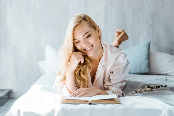 Bela menina loira feliz em pijama deitado na cama e livro de leitura — Fotografia de Stock