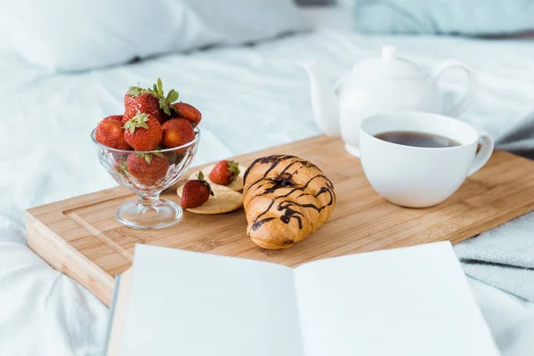 Аппетитный завтрак из клубники, круассана и кофе на деревянном подносе на кровати — стоковое фото