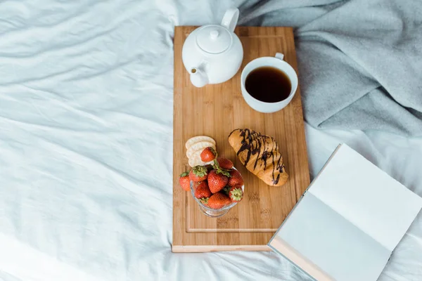 Vista de alto ângulo de pequeno-almoço apetitoso de morangos, croissant e café na bandeja de madeira na cama — Fotografia de Stock