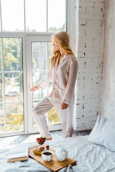 Mulher loira de pijama abrindo janela, café da manhã e livro na cama de manhã — Fotografia de Stock