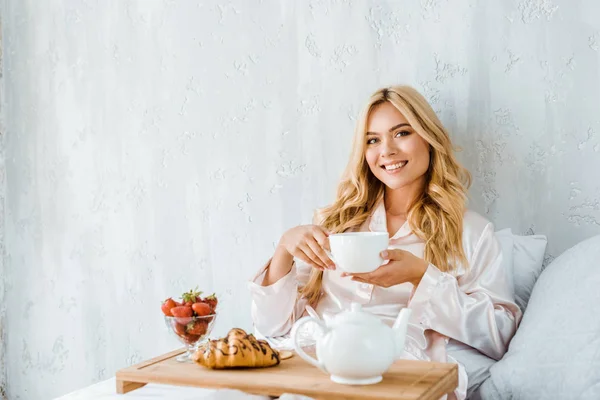 Schöne lächelnde Frau, die morgens eine Tasse Tee im Bett hält und in die Kamera schaut — Stockfoto