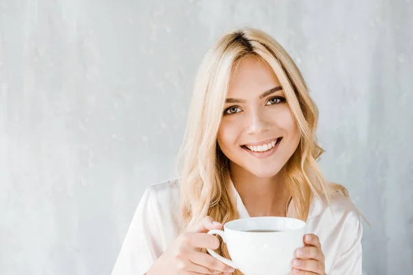 Ritratto di donna attraente in pigiama che tiene in mano una tazza di tè e guarda la fotocamera in camera da letto — Foto stock