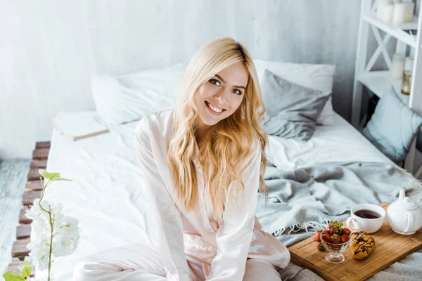Mujer atractiva sonriente en pijama mirando a la cámara, desayuno en bandeja de madera en la cama - foto de stock