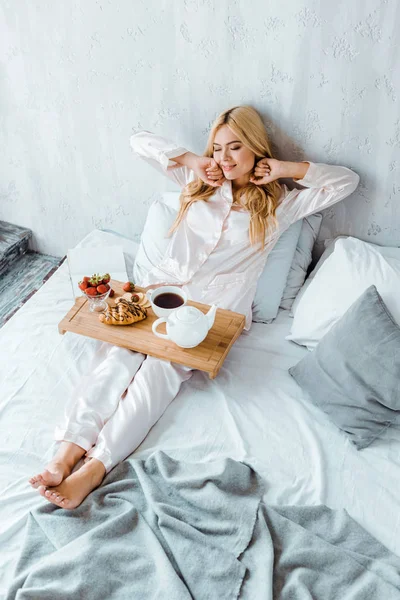 Vista de ángulo alto de mujer atractiva en pijama estirándose en la cama y sosteniendo bandeja de madera con desayuno - foto de stock