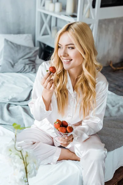 Atractiva mujer sonriente en pijama comiendo fresas en la cama por la mañana y mirando hacia otro lado - foto de stock