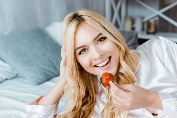 Lächelnde attraktive Frau im Schlafanzug, die morgens Erdbeere im Bett isst und in die Kamera schaut — Stockfoto