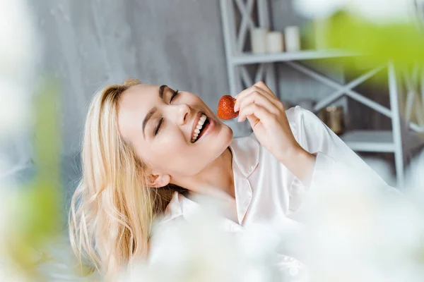 Atractiva mujer en pijama comiendo fresa en la cama por la mañana con los ojos cerrados - foto de stock